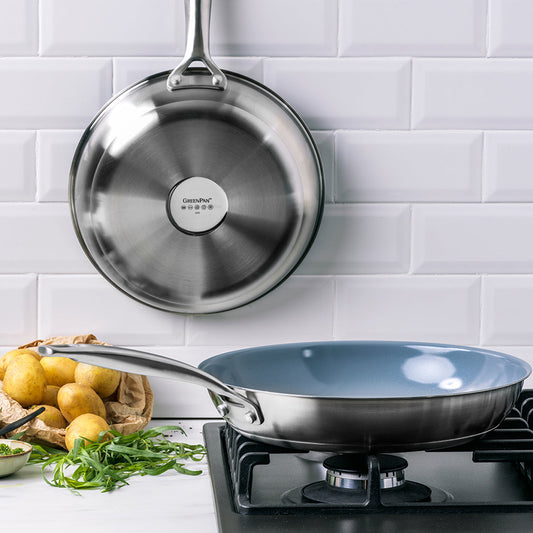 Two stainless steel nonstick frypans on a stove, one searing meat, with fresh herbs and potatoes nearby.