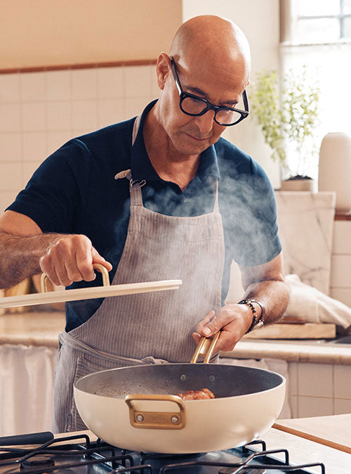 Man with glasses and apron lifting lid from a steaming white ceramic nonstick skillet in a modern kitchen.