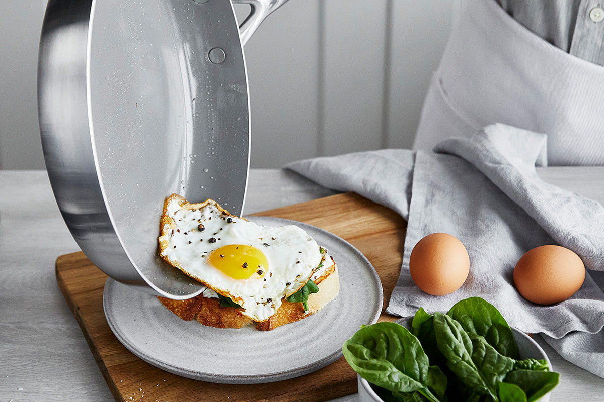 Ceramic nonstick skillet sliding a fried egg onto toast, with whole eggs and spinach on a wooden surface.
