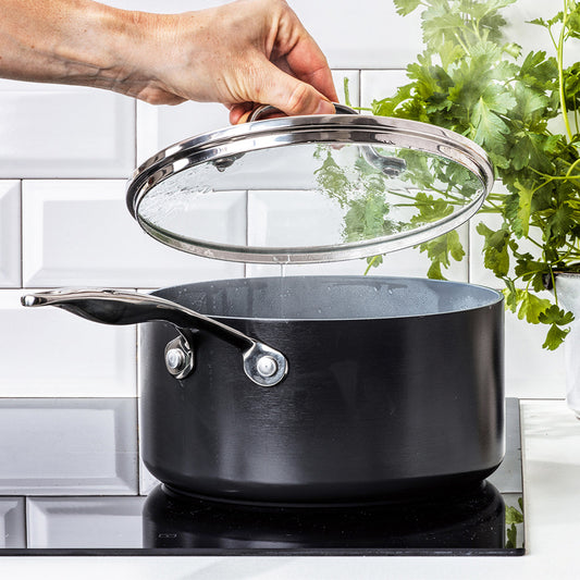 Hand lifting lid from steaming 2-quart ceramic nonstick saucepan on stove, fresh herbs visible in the background.