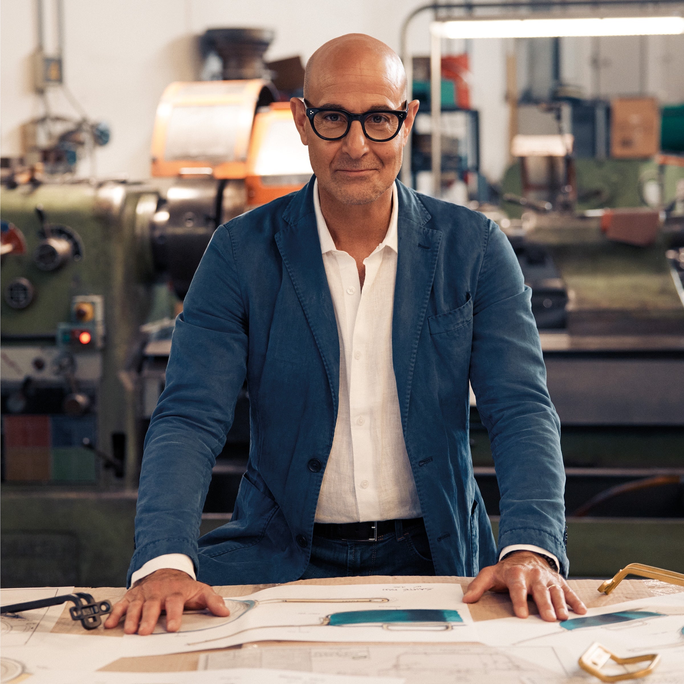 Stanley Tucci standing at a table with blueprints in an industrial setting