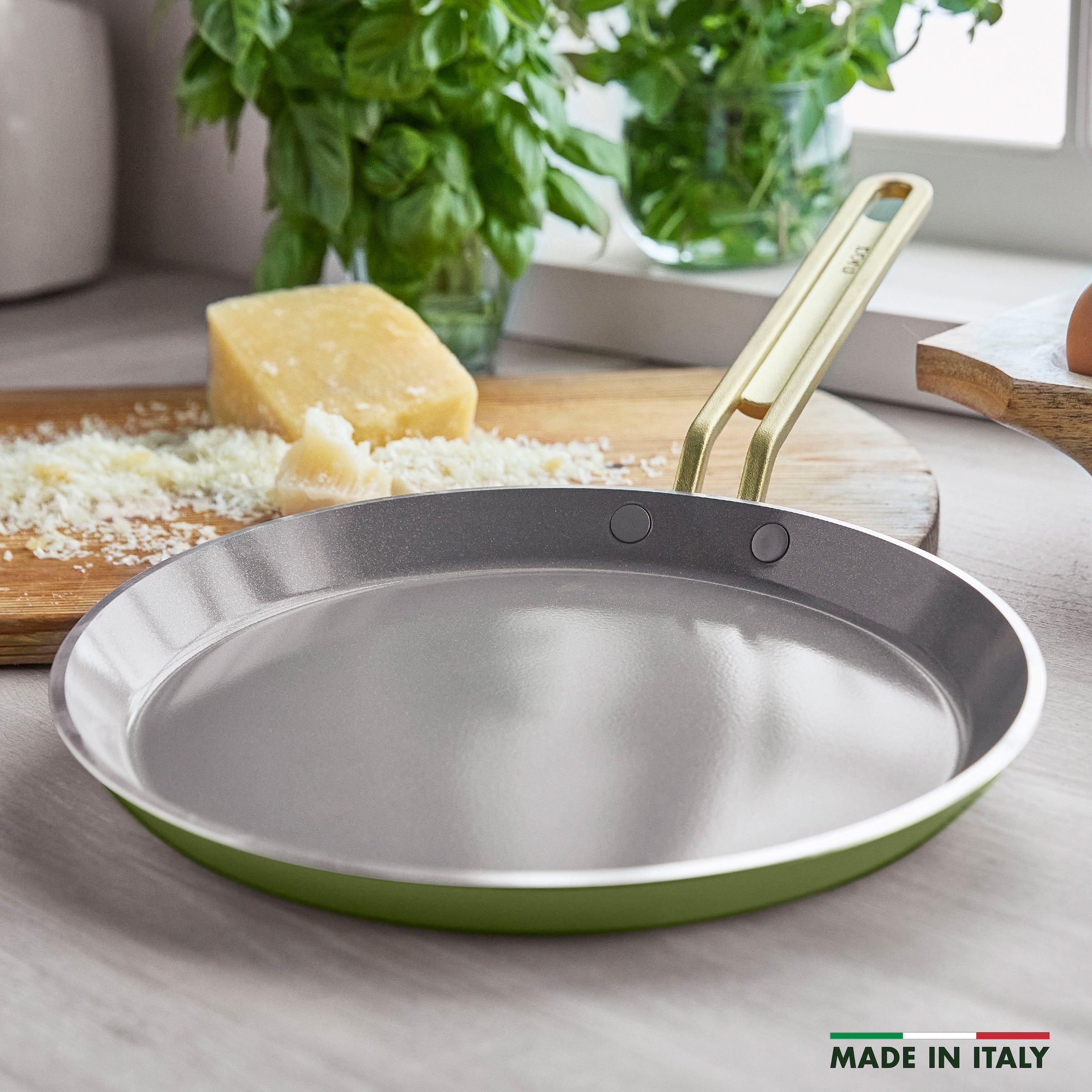 Green crepe pan on a kitchen counter with cheese and herbs in the background.
