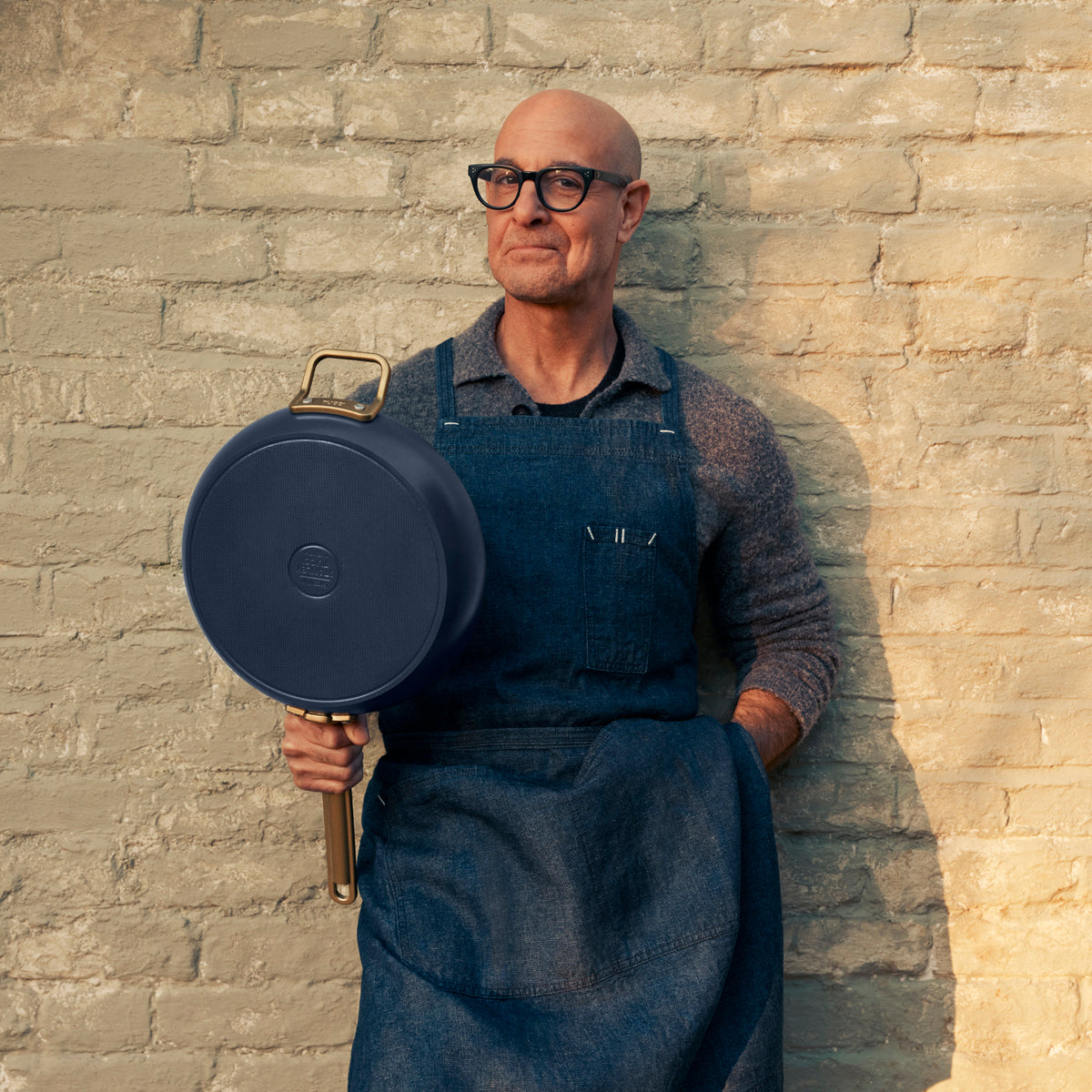Stanley Tucci holding a blue saute pan against a brick background